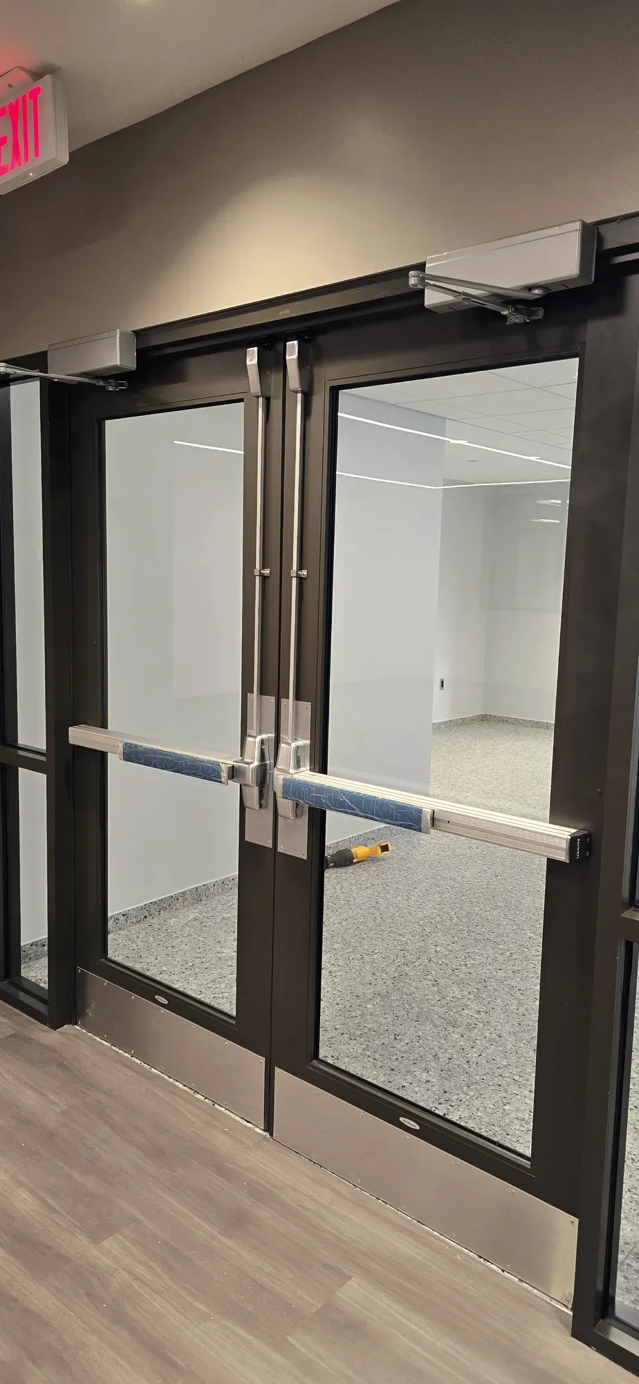 Vertical-rod panic bars and closers on dark-framed glass storefront double doors